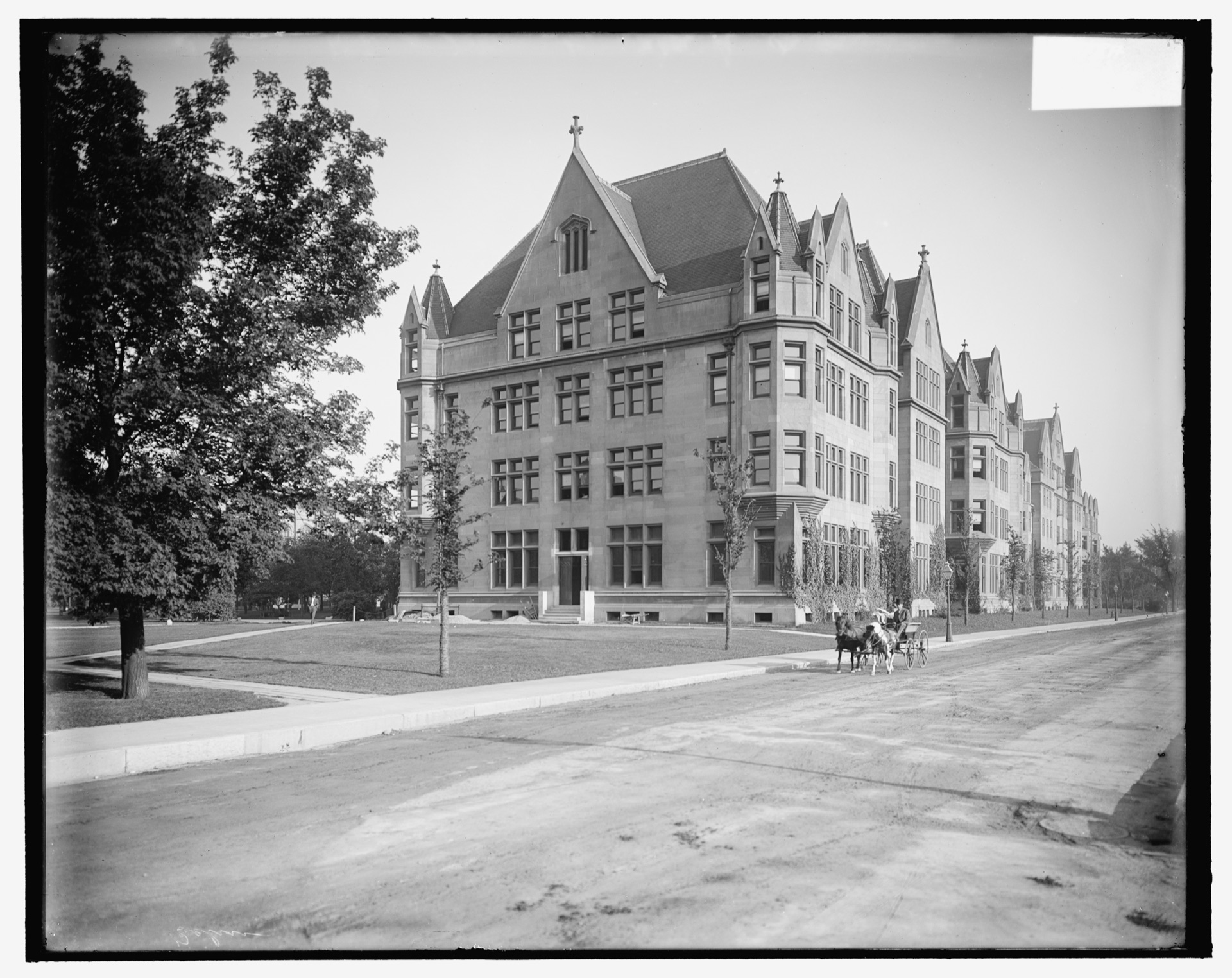 Cobb Lecture Hall about 1900, a long four-storey limestone Gothic block with steep gables and turrets, young trees along the sidewalk and a horse and buggy passing on the unpaved street
