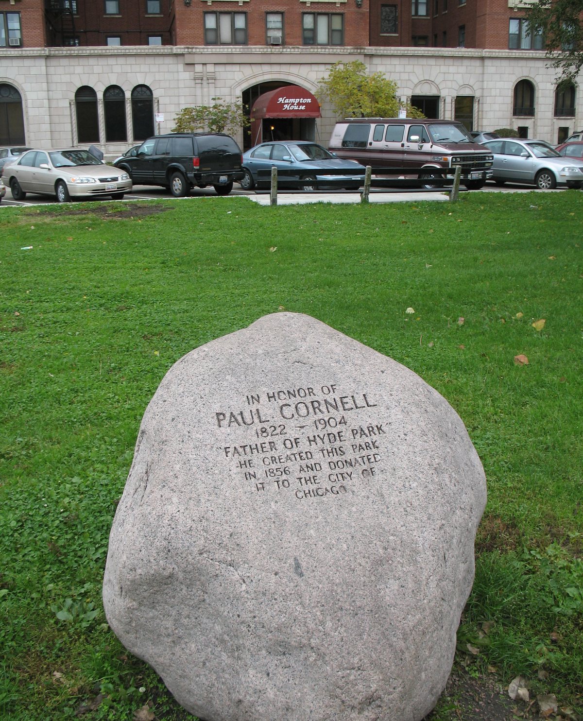 A granite boulder on a lawn carved with the words IN HONOR OF PAUL CORNELL 1822 to 1904, FATHER OF HYDE PARK, HE CREATED THIS PARK IN 1856 AND DONATED IT TO THE CITY OF CHICAGO, with the Hampton House behind it across a row of parked cars
